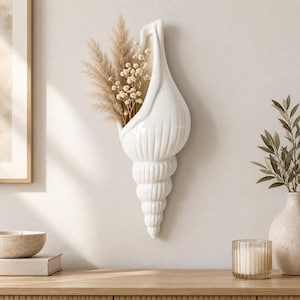 Puede incluir: Un florero de pared de cerámica blanca con forma de concha marina en espiral, con un acabado brillante y crestas verticales. El florero contiene un arreglo decorativo de hierba de la pampa beige seca y pequeñas flores blancas.