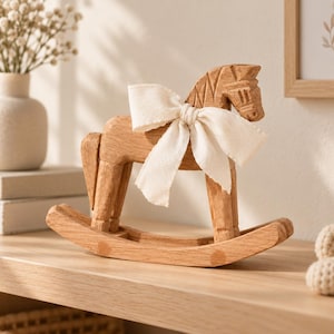 May include: A carved wooden rocking horse figurine featuring a light brown natural wood grain finish. The horse is adorned with a tied off-white fabric ribbon around its neck, showcasing detailed etched lines on the mane and face.