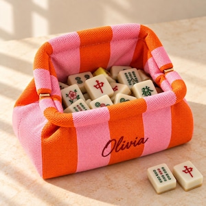 Può includere: Una pochette in maglia a righe verticali arancioni e rosa con il nome Olivia ricamato in corsivo rosso scuro. La pochette è riempita con tessere del mahjong color crema, e due tessere aggiuntive sono appoggiate all'esterno.