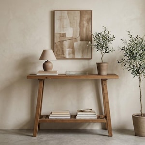 May include: A wooden console table with a lower slatted shelf, topped with a beige lamp, a stack of books, and a small tray. Above the table hangs a framed abstract painting in shades of beige and brown, next to a potted olive tree.