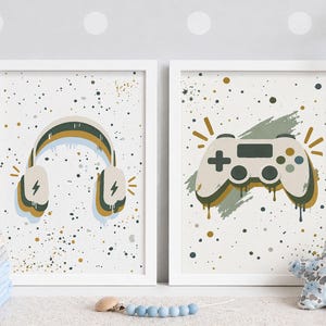 May include: Two framed wall art prints featuring a white background with multicolored paint splatters. The left print displays a pair of headphones with a lightning bolt icon, while the right print shows a video game controller with a brush stroke.