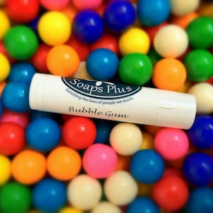 May include: A white tube of lip balm with the words "Soaps Plus" and "Bubble Gum" printed on it. The tube is surrounded by colorful gumballs.