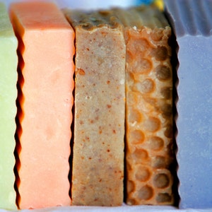 May include: A set of six bars of handmade soap in various colours, including green, orange, brown, yellow, purple, and blue. The bars are arranged in a row, with the green bar on the left and the blue bar on the right.