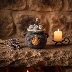 May include: A dark gray crocheted cauldron featuring two side handles, a green rim, and a felt flame appliqué on the front. The top is filled with crocheted gray spheres and a small white sphere, resembling a bubbling liquid inside the pot.