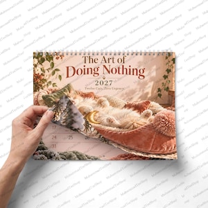 May include: A 2027 wall calendar titled The Art of Doing Nothing, Twelve Cats, Zero Urgency. The cover features a ginger cat sleeping in a plush, dusty rose-colored bed with a pom-pom trim. A hand is shown turning the spiral-bound page.