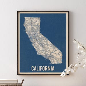 May include: A framed wall art print featuring a detailed map of California in beige against a deep blue background. The map displays intricate road networks and geographical features. The word CALIFORNIA is printed in bold white capital letters.