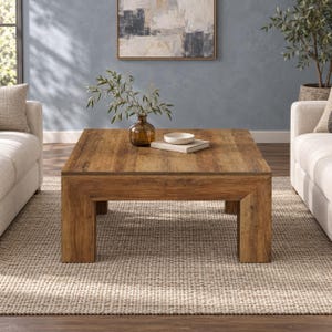 Könnte beinhalten: Ein quadratischer Couchtisch mit einer mittelbraunen Holzmaserung und dicken, stabilen Beinen. Auf der Tischoberfläche steht eine klare Glasvase mit grünen Blattzweigen sowie eine kleine weiße Keramikschale auf einem Stapel von zwei Büchern.