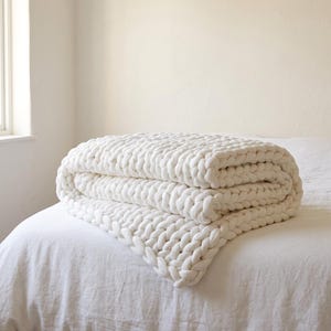 May include: A chunky knit blanket in a solid off-white color, featuring large, thick yarn loops woven into a consistent, heavy braided pattern. The blanket is folded into three distinct layers, showcasing the texture of the oversized knit stitches.