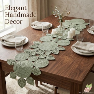 Puede incluir: Un camino de mesa de ganchillo en color verde salvia que presenta una serie de motivos circulares y en forma de hoja interconectados. El camino está colocado sobre una mesa de madera con platos blancos, servilletas y velas.