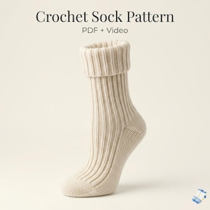 May include: A cream-colored crochet sock with a ribbed texture and a folded cuff. The text at the top reads Crochet Sock Pattern PDF + Video. The sock features a vertical knit-like stitch pattern and a rounded toe and heel construction.