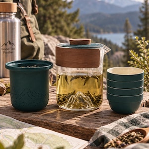 Pode incluir: Um bule de vidro transparente com tampa de madeira e base em forma de montanha, um infusor de chá verde-azulado escuro com um padrão de montanha pontilhado e uma pilha de quatro chávenas de chá a condizer em verde-azulado escuro.