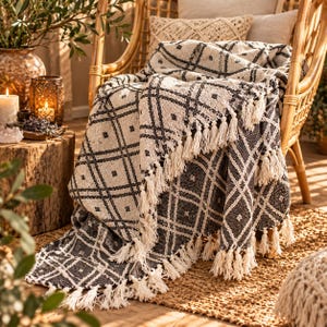 May include: A woven throw blanket featuring a black and cream diamond grid pattern with small square accents. The blanket has a textured surface and is finished with long, cream-coloured fringe tassels along the edges, draped over a rattan chair.