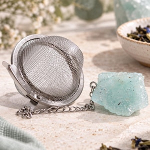 Può includere: Un infusore per tè a sfera in rete di acciaio inossidabile con chiusura a cerniera. Una catenella in metallo argentato collega l'infusore a una pietra di cristallo grezza color verde menta pallido. La rete è fine e circolare, per tè sfuso.