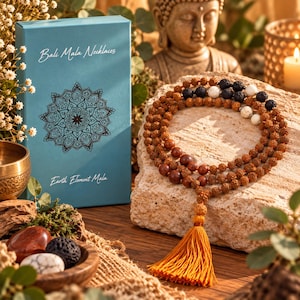Pode incluir: Um colar mala com contas de rudraksha castanhas, pedras de lava pretas e contas de mármore branco, finalizado com uma borla laranja. O colar é exibido ao lado de uma caixa azul com a etiqueta Bali Mala Necklaces e Earth Element Mala.