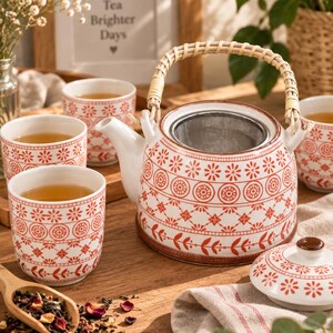 Op de afbeelding: Een witte keramische theepot en drie bijpassende kopjes met een rood bloemen- en geometrisch patroon. De theepot heeft een gebogen tuit, een uitneembare metalen zeef en een lichtgekleurd handvat van gevlochten bamboe.