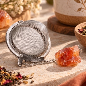 Peut inclure: Une boule à thé sphérique en acier inoxydable avec un maillage fin et un loquet métallique marqué SUS 304. Une chaîne argentée relie l'infuseur à un cristal brut orange-rouge translucide qui sert de poignée décorative.