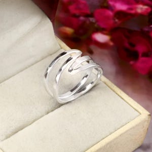 Puede incluir: Un anillo de plata pulida con un diseño calado de varias capas, compuesto por tres bandas lisas y distintas que se curvan y se superponen para crear una forma fluida y continua. El anillo tiene un acabado metálico brillante y un aspecto elegante.