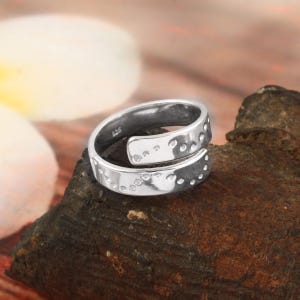 May include: A silver-coloured bypass ring featuring a polished finish and a series of small, circular indentations along the edges. The inner band is clearly stamped with the hallmark 925, indicating the metal composition of this open-style ring.
