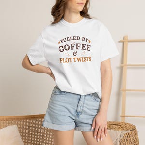 Puede incluir: Camiseta blanca con el texto "FUELED BY COFFEE & PLOT TWISTS" en letras marrones y naranjas. La camiseta se lleva con pantalones cortos vaqueros. El tema general es la ropa informal.