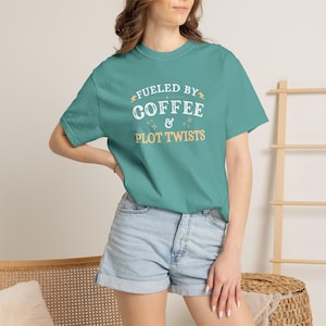 Puede incluir: Camiseta de color turquesa con el texto "Fueled by Coffee & Plot Twists" en letras blancas y doradas. La camiseta tiene cuello redondo y mangas cortas. La modelo lleva pantalones cortos vaqueros.