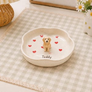 Puede incluir: Un plato pequeño redondo de cerámica color hueso con borde festoneado, que presenta una pequeña figura tridimensional de un golden retriever en el centro. El plato está decorado con varios corazones rojos pequeños y el nombre Teddy en negro.