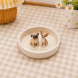 Puede incluir: Un plato decorativo redondo de cerámica color crema con dos pequeñas figuras de perro, un pug y un chihuahua, fijadas en el centro. El texto "Buddy & Lucy" está grabado en la base del plato, que tiene un acabado liso y mate.