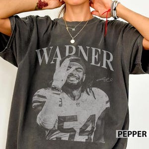 May include: A charcoal grey short-sleeve t-shirt featuring a faded black and white graphic of a football player wearing a jersey with the number 54. The word WARNER is printed in large capital letters above the player, and the word PEPPER is visible.