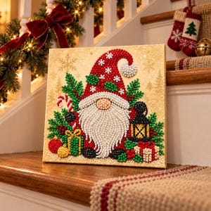 May include: A square diamond painting canvas featuring a gnome with a long white beard, a red hat with white snowflakes, and a pom-pom. The gnome is surrounded by green pine branches, red berries, a candy cane, two wrapped gifts, and a black lantern.