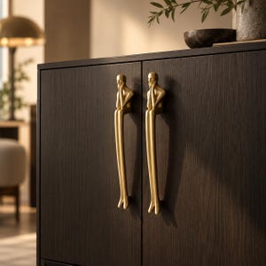 Peut inclure: Deux poignées de placard en métal doré montées sur une porte de placard en bois brun foncé à grain apparent. Chaque poignée représente une silhouette humaine stylisée en position assise, aux jambes allongées et aux mains près du visage.