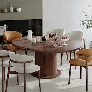 May include: A round dark wood dining table with a central fluted pedestal base, surrounded by six chairs featuring curved wooden frames and upholstered seats in alternating cream and tan colors, with a wine bottle and glassware on top.