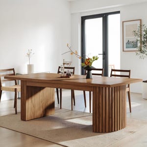 May include: A rectangular wooden dining table featuring a smooth top and two distinct bases: one flat panel support and one semi-circular base with vertical slats. The table is surrounded by four wooden chairs with woven seats.