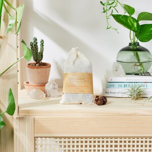 May include: A white cloth bag filled with organic soap nuts, labeled "BUTTER ME UP ORGANICS Organic Soap Nuts 4 oz (113 g) - Makes 75 Loads". The bag is sitting on a wooden shelf with a potted cactus, crystals, and books.