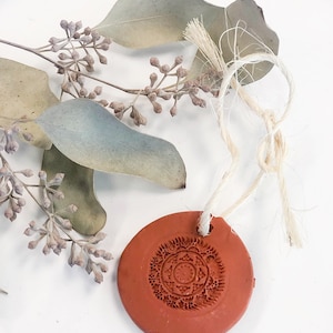 Puede incluir: Un adorno de arcilla de terracota con un diseño de mandala detallado, colgado de una cuerda de hilo. El adorno está sobre una superficie blanca con hojas de eucalipto secas.