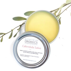May include: A silver tin with a yellow lid containing Calendula Salve by Butter Me Up Organics. The label reads "Butter Me Up Organics, a modern day apothecary, Calendula Salve, 2 oz. Apply as needed to wounds, rashes, bites, and broken skin for optimum and rapid healing. Ingredients: Calendula Flowers, Olive Oil, Shea Butter* *Indicates certified organic www.butterneuporganics.com"