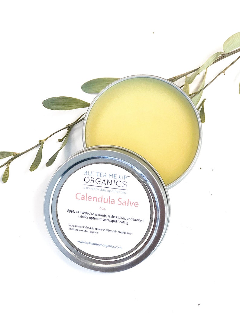 May include: A silver tin with a yellow lid containing Calendula Salve by Butter Me Up Organics. The label reads "Butter Me Up Organics, a modern day apothecary, Calendula Salve, 2 oz. Apply as needed to wounds, rashes, bites, and broken skin for optimum and rapid healing. Ingredients: Calendula Flowers, Olive Oil, Shea Butter* *Indicates certified organic www.butterneuporganics.com"