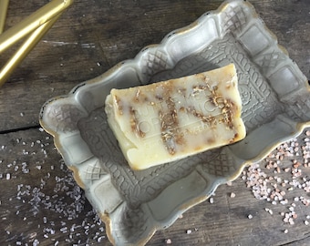 Puede incluir: Una barra de jabón en una bandeja decorativa. El jabón es de color amarillo claro y tiene un remolino marrón. El jabón está etiquetado como "Butternut Organics".