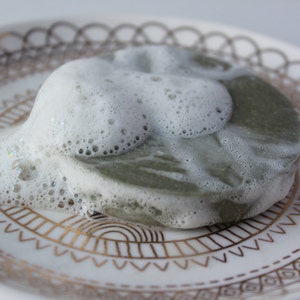 Puede incluir: Una pastilla de jab&oacute;n verde cubierta de espuma blanca sobre un plato blanco con un patr&oacute;n dorado.