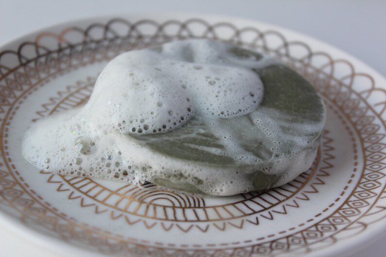 Puede incluir: Una pastilla de jab&oacute;n verde cubierta de espuma blanca sobre un plato blanco con un patr&oacute;n dorado.