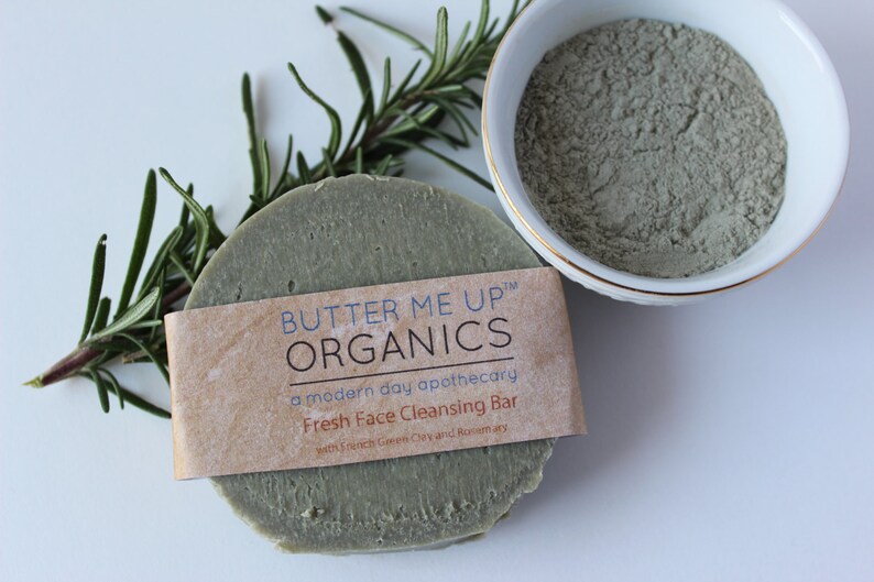 Puede incluir: Una barra redonda de jab&oacute;n verde con una etiqueta marr&oacute;n que dice "BUTTER ME UP&trade; ORGANICS a modern day apothecary Fresh Face Cleansing Bar with French Green Clay and Rosemary".