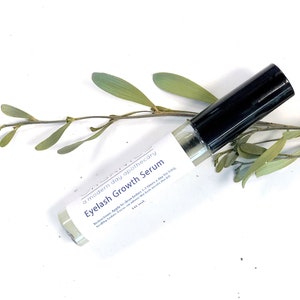 May include: A clear glass bottle of Eyelash Growth Serum with a black cap, labeled with the text "Eyelash Growth Serum" and "a modern day apothecary". The bottle is 10 ml and is set against a white background with green leaves.
