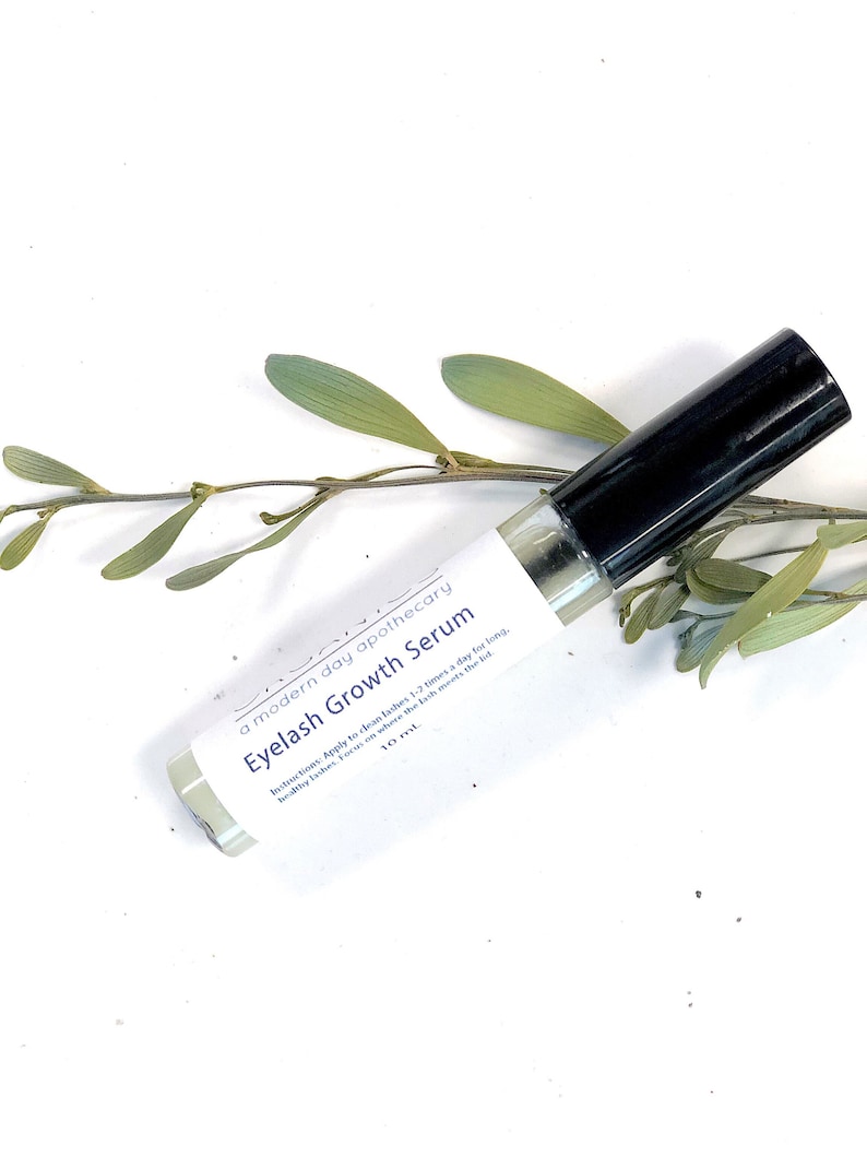 May include: A clear glass bottle of Eyelash Growth Serum with a black cap, labeled with the text "Eyelash Growth Serum" and "a modern day apothecary". The bottle is 10 ml and is set against a white background with green leaves.