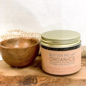 May include: A glass jar of charcoal toothpaste with a gold lid. The jar is labeled "Butter Me Up Organics" and "Charcoal Toothpaste with Activated Charcoal". The jar is sitting on a wooden surface.