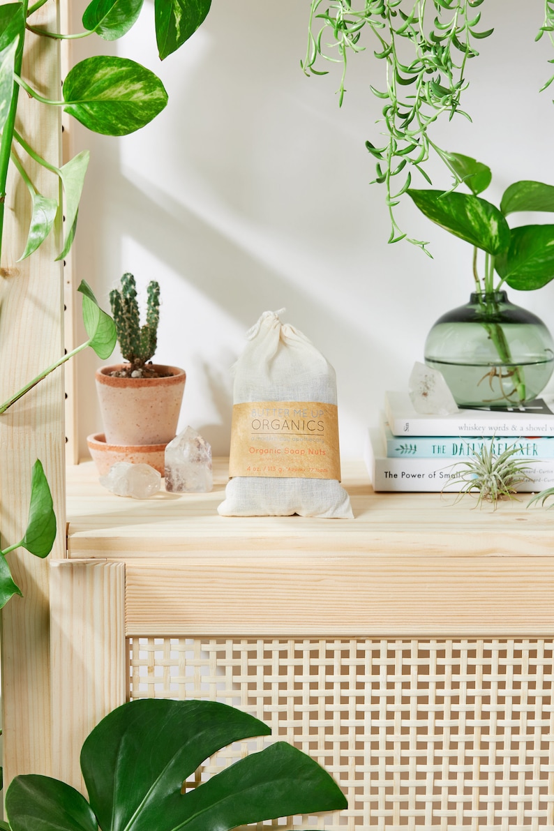 May include: A white cloth bag with a brown label that reads "Butter Me Up Organics" and "Organic Soap Nuts". The bag is sitting on a wooden shelf with a cactus, crystals, and books.