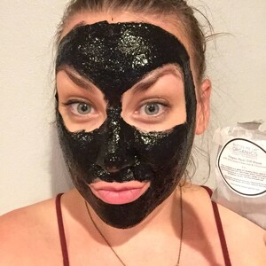 May include: A woman with a black peel-off face mask on. The mask is covering her entire face except for her eyes and eyebrows. She is holding a small bag with the text "Butter Me Up Organics Vegan Peel Off Mask with Activated Charcoal & Green Tea" on it.