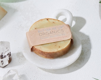 May include: A white marble soap dish with a round, beige soap bar wrapped in brown paper. The soap bar is labeled "Butter Me Up Organics" and "Healing Face Cleansing Bar with Turmeric & Calendula".