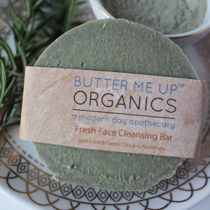 Puede incluir: Una barra de jab&oacute;n redonda verde con una etiqueta que dice "Butter Me Up Organics, una botica moderna, Barra limpiadora facial fresca con arcilla verde francesa y romero".