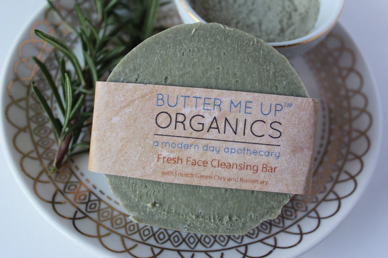 Puede incluir: Una barra de jab&oacute;n redonda verde con una etiqueta que dice "Butter Me Up Organics, una botica moderna, Barra limpiadora facial fresca con arcilla verde francesa y romero".
