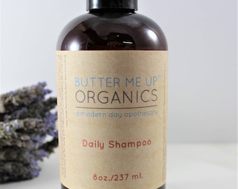 May include: A brown glass bottle with a black cap and a brown paper label. The label says "Butter Me Up Organics, a modern day apothecary, Daily Shampoo, 8oz/237 ml."