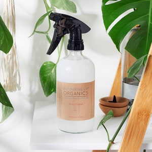 May include: A clear glass bottle with a black spray nozzle filled with a white liquid. The bottle has a brown label with the text "Butter Me Up Organics" and "Glass Cleaner" on it. The bottle is sitting on a white shelf with green plants in the background.