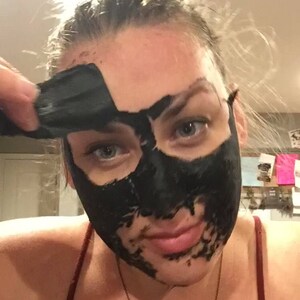 May include: A woman is applying a black charcoal face mask to her face. The mask is already applied to her forehead, nose, and chin.